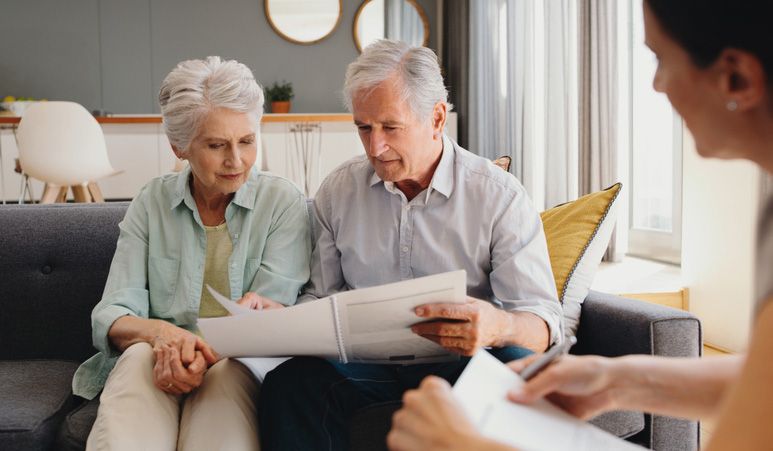 Elderly couple reviewing paperwork with a younger person, seated on a couch indoors.
