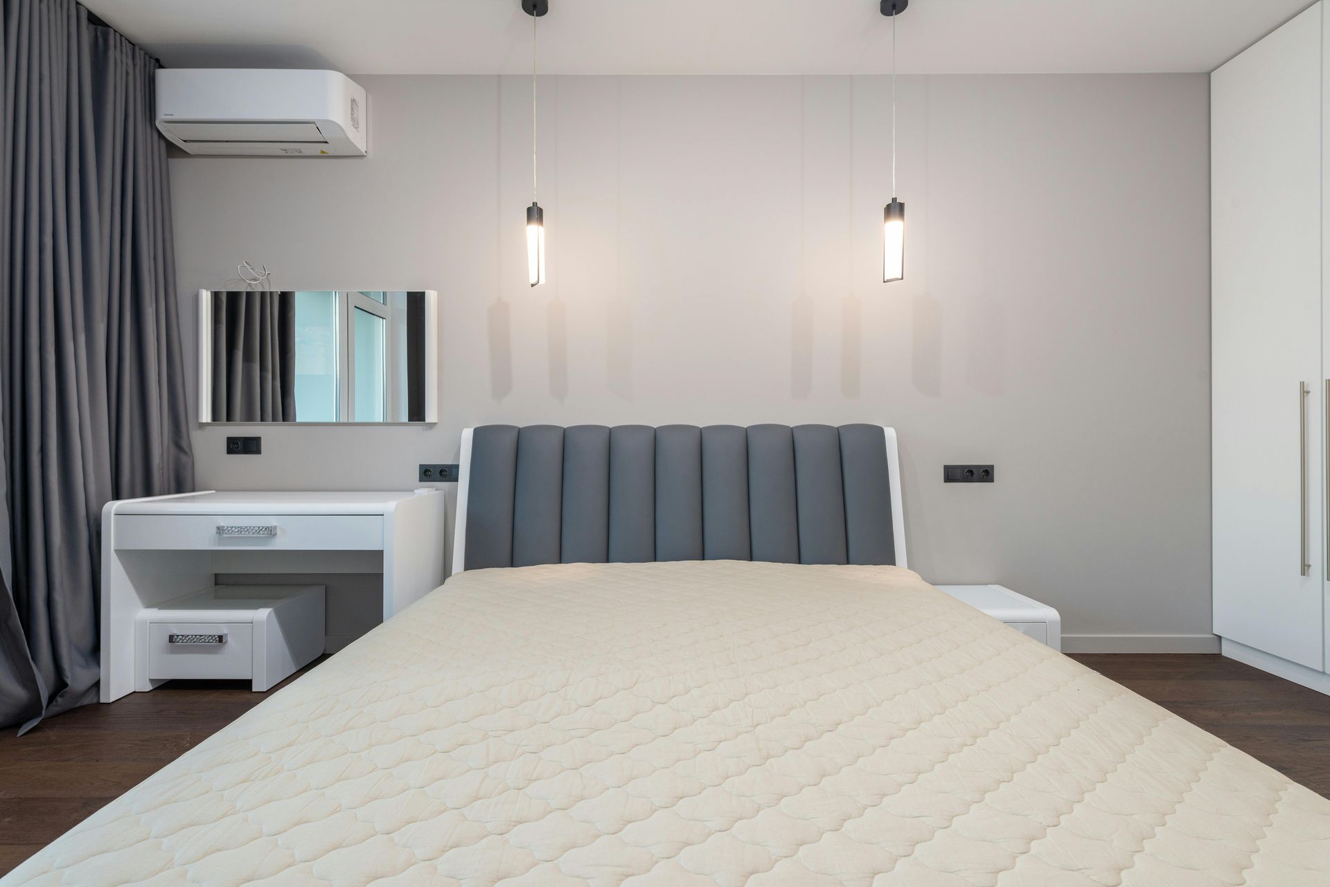 A bedroom with a beige mattress, a gray upholstered headboard, a white vanity table, and gray curtains.