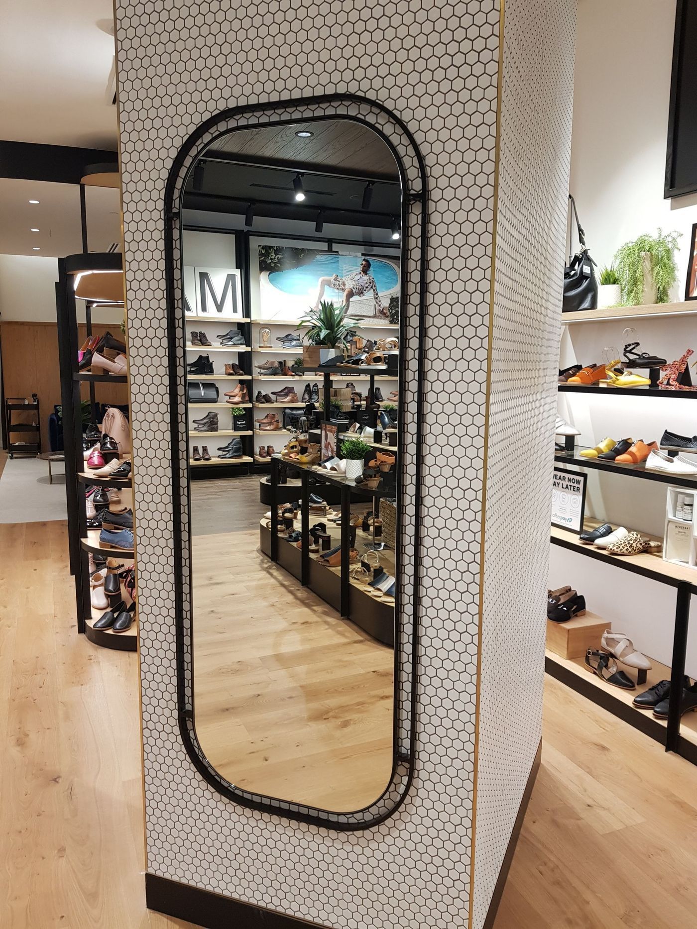 Full-length mirror in a shoe store. White wall with black dot pattern, black frame, wooden floor, shoe displays.