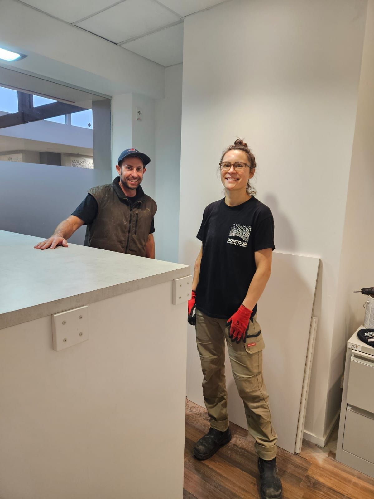 Two people smiling in a room. A man leans on a white counter, woman stands with tools, wearing work clothes.