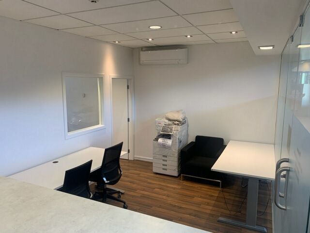 Office with white walls, wooden floor, two desks, chair, couch, copier, window, and AC unit.