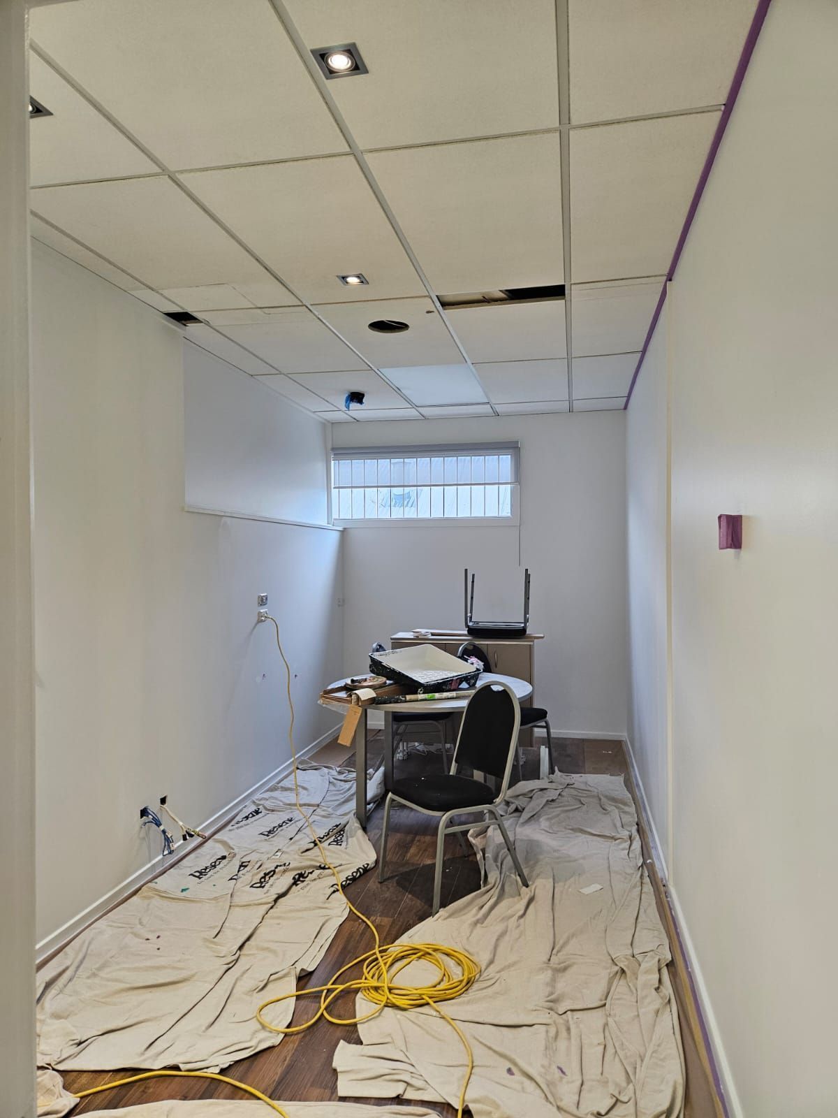 A small office under renovation with paint tarps, exposed ceiling tiles, a desk, and a window.
