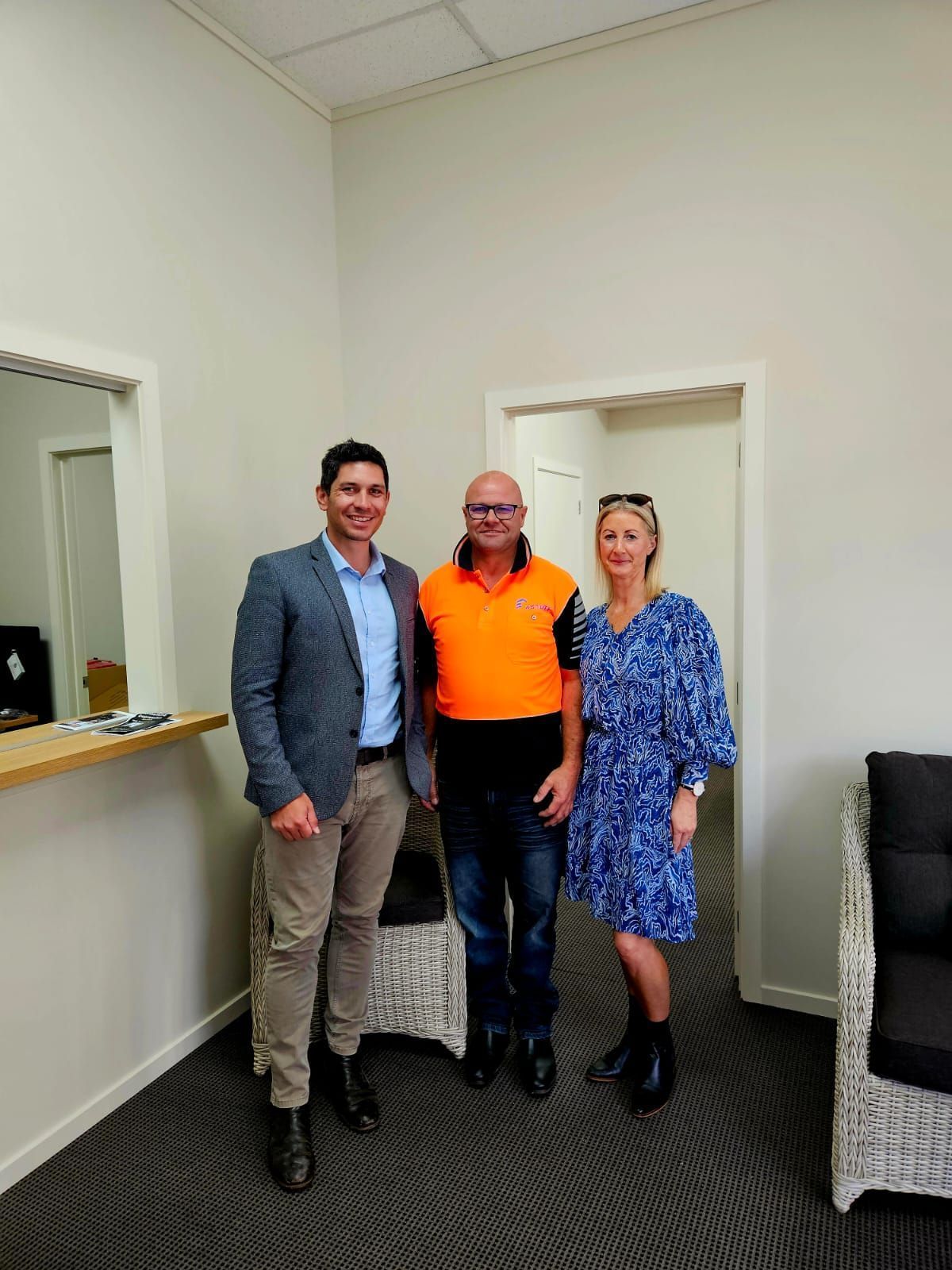 Three people stand in an office. Man in a blazer, man in orange shirt, woman in a blue dress, smiling.