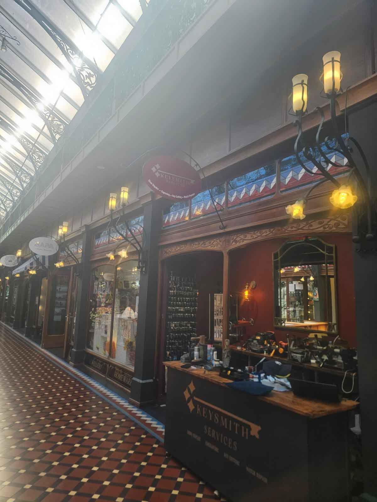 Interior of a vintage shopping arcade with shops on both sides, glass ceiling, and red tile floor.