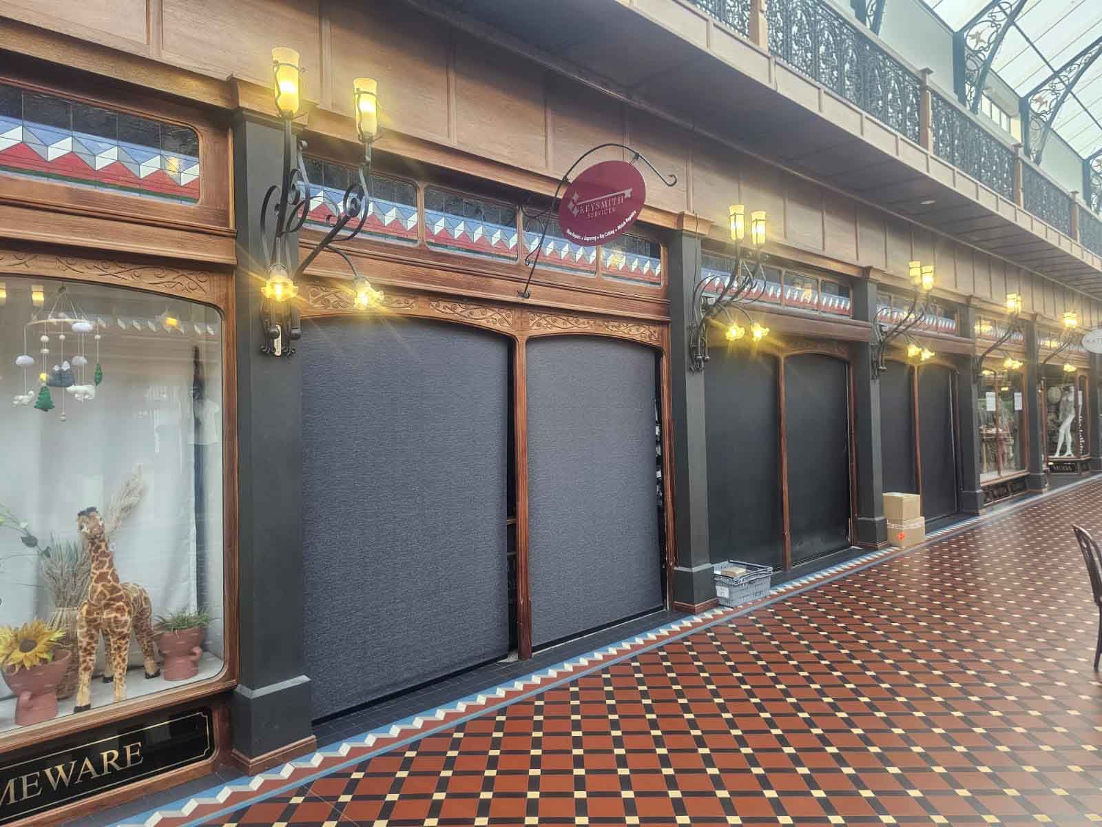 Shuttered shopfronts in a brick-paved arcade with a decorative overhead walkway.