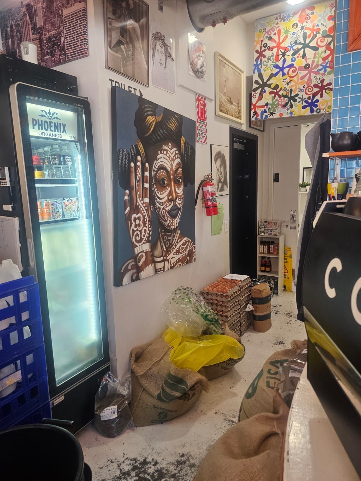 Cafe interior: Art, cooler, bags of coffee beans on floor, open doorway, and a black espresso machine.