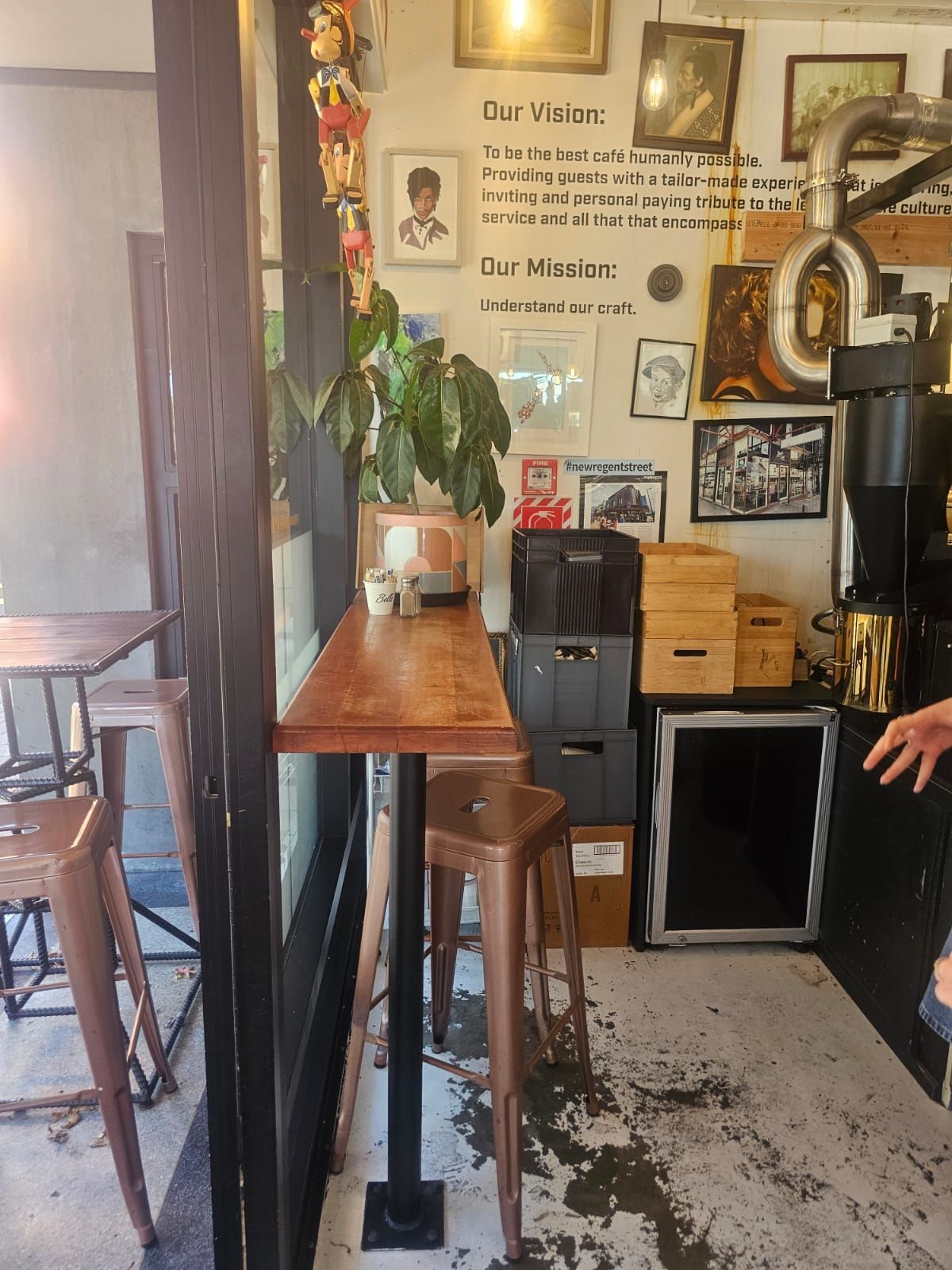 A cafe interior with a wooden counter, stools, plants, and framed art on the wall.