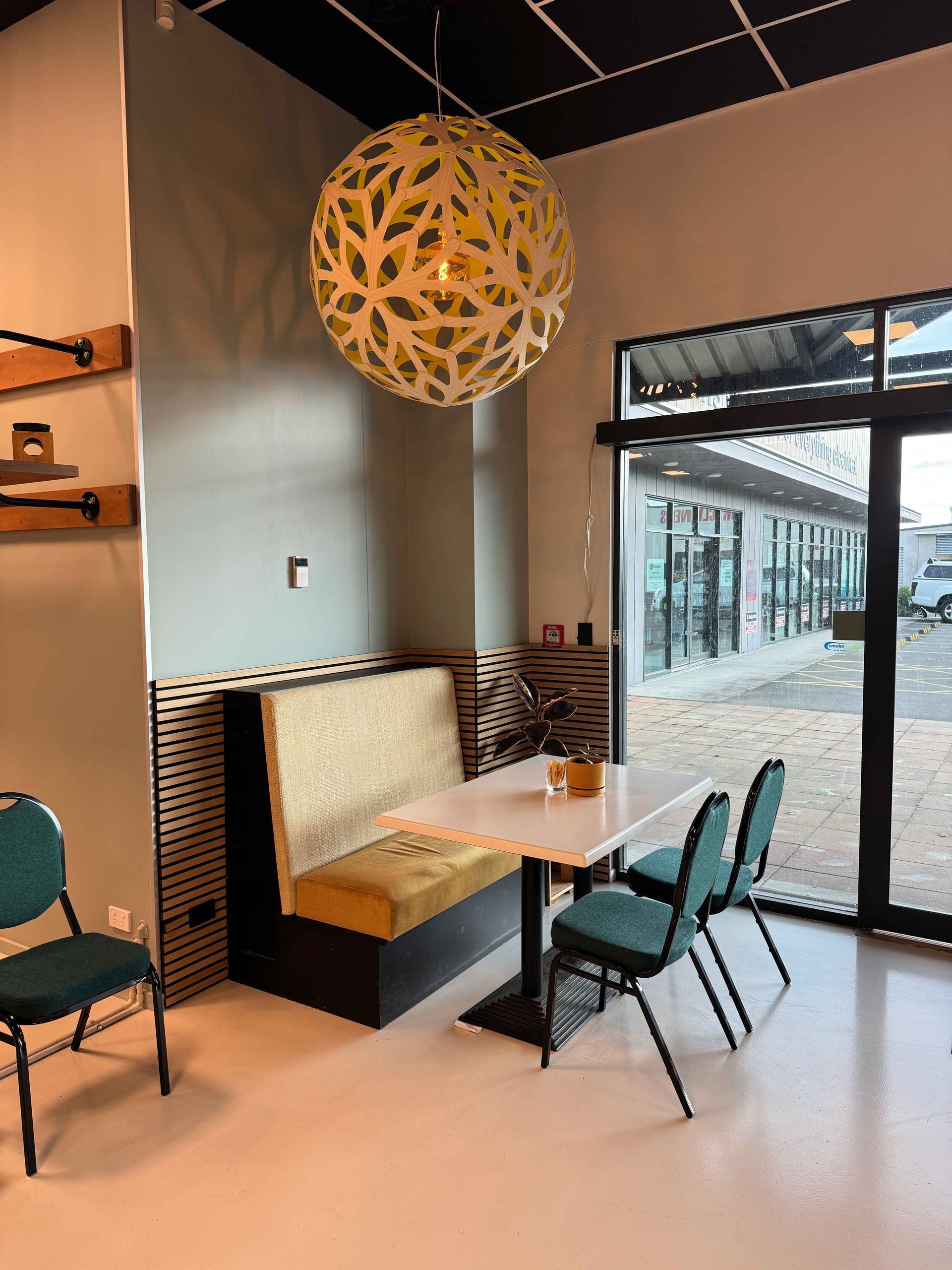 Cozy cafe corner: booth with gold cushion, small table, green chairs, patterned globe light, and a large glass door.