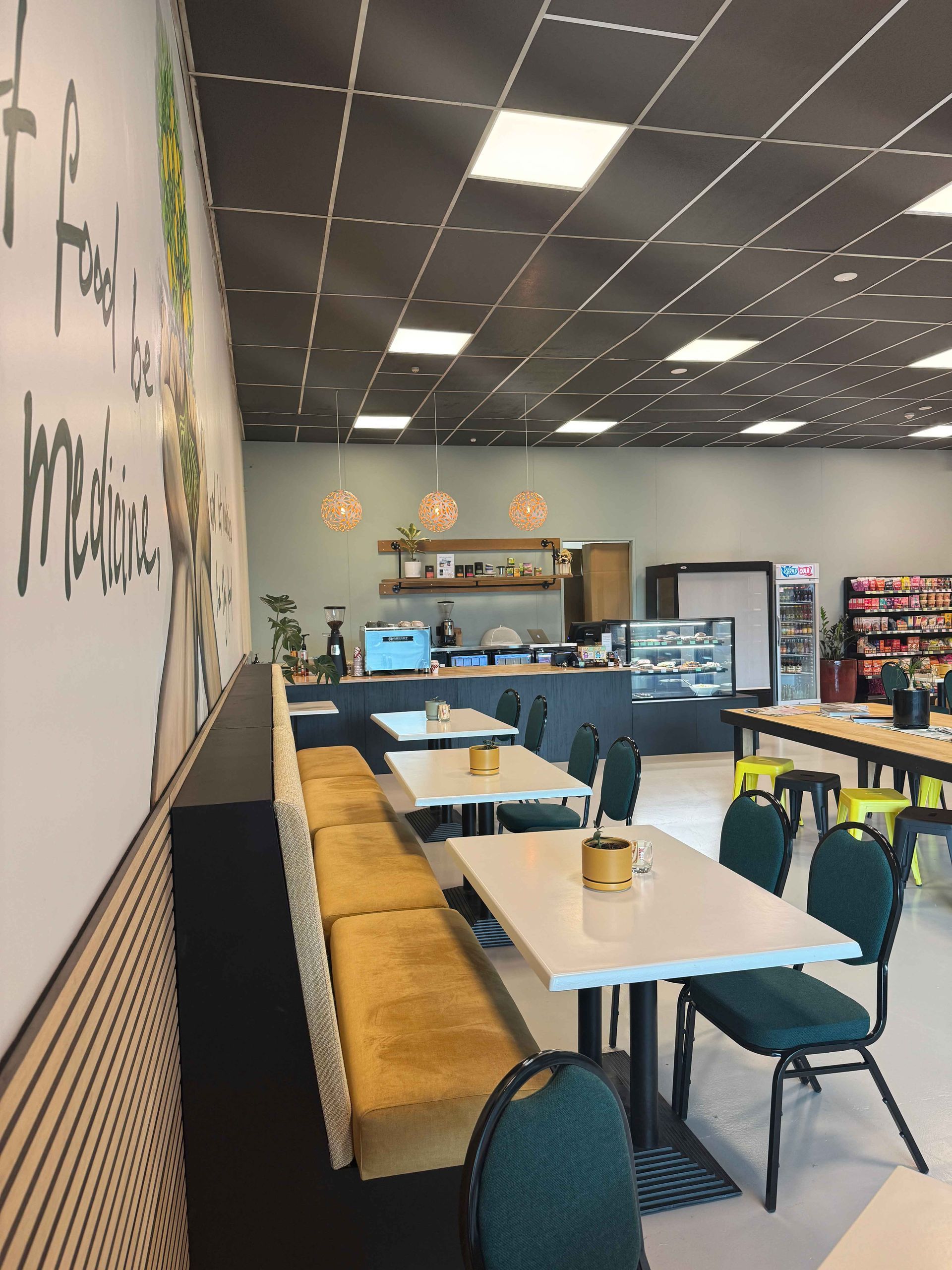 Cafe interior with tables, a bench, and a counter. Walls are white and gray. Some decor and a fridge visible.