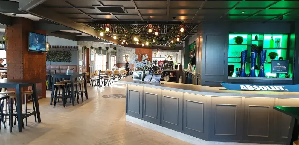 Interior of a bar with a long counter and tables. Green shelving highlights liquor.