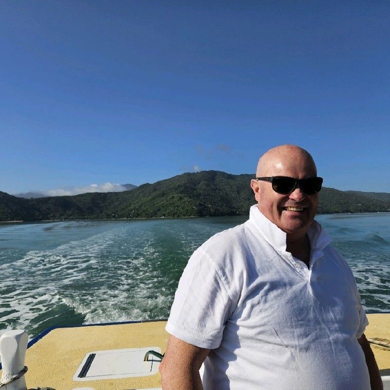 Man in sunglasses on a boat, smiling, with mountains and blue sky in the background.
