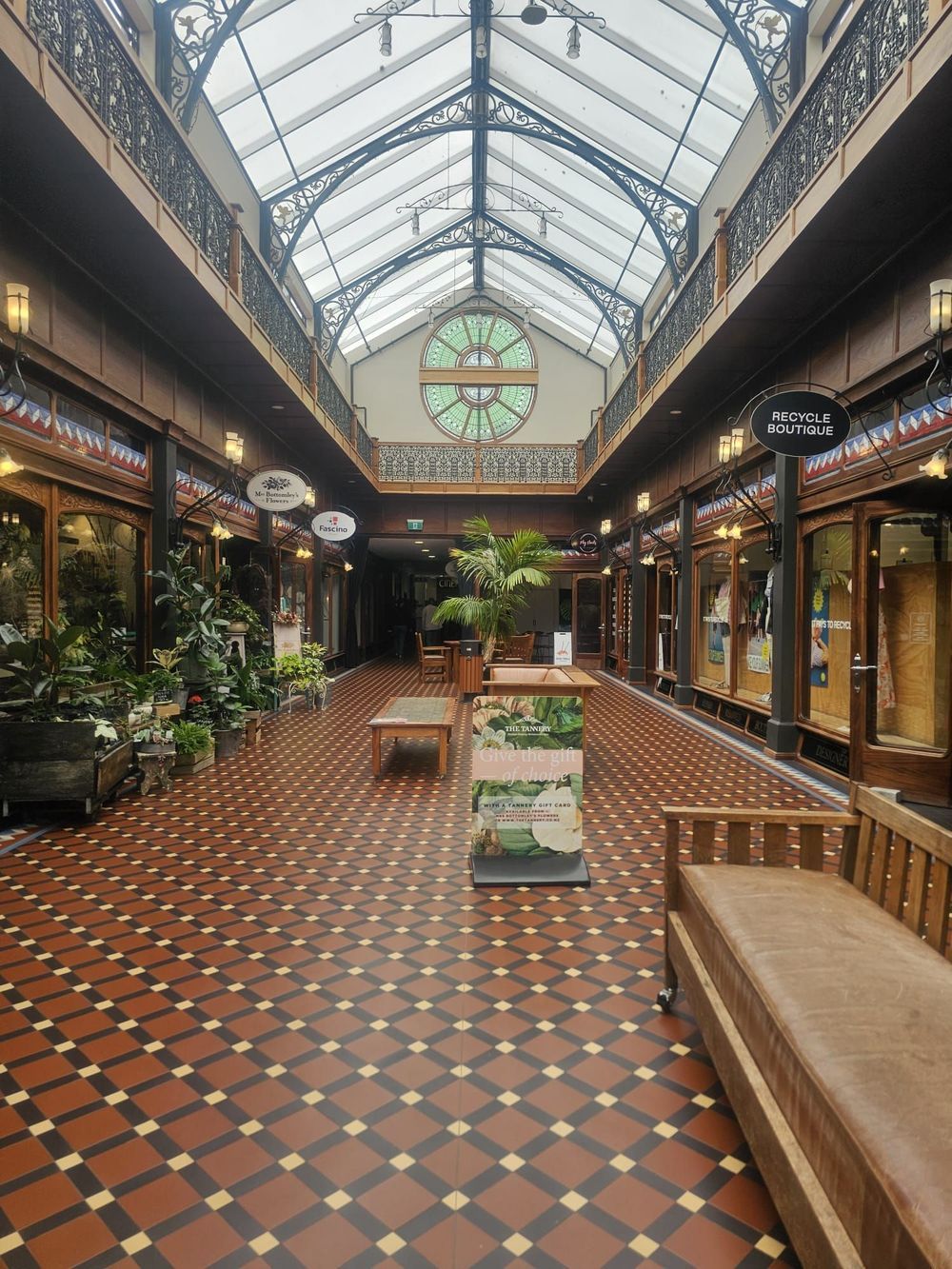 Long, covered arcade with shops, red tile floor, glass ceiling, plants, benches.