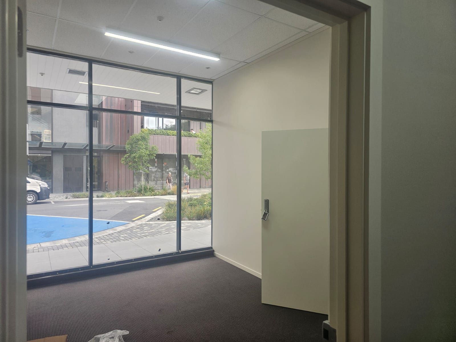 Empty room with large window overlooking an exterior space with a building, trees, and a blue paved area.