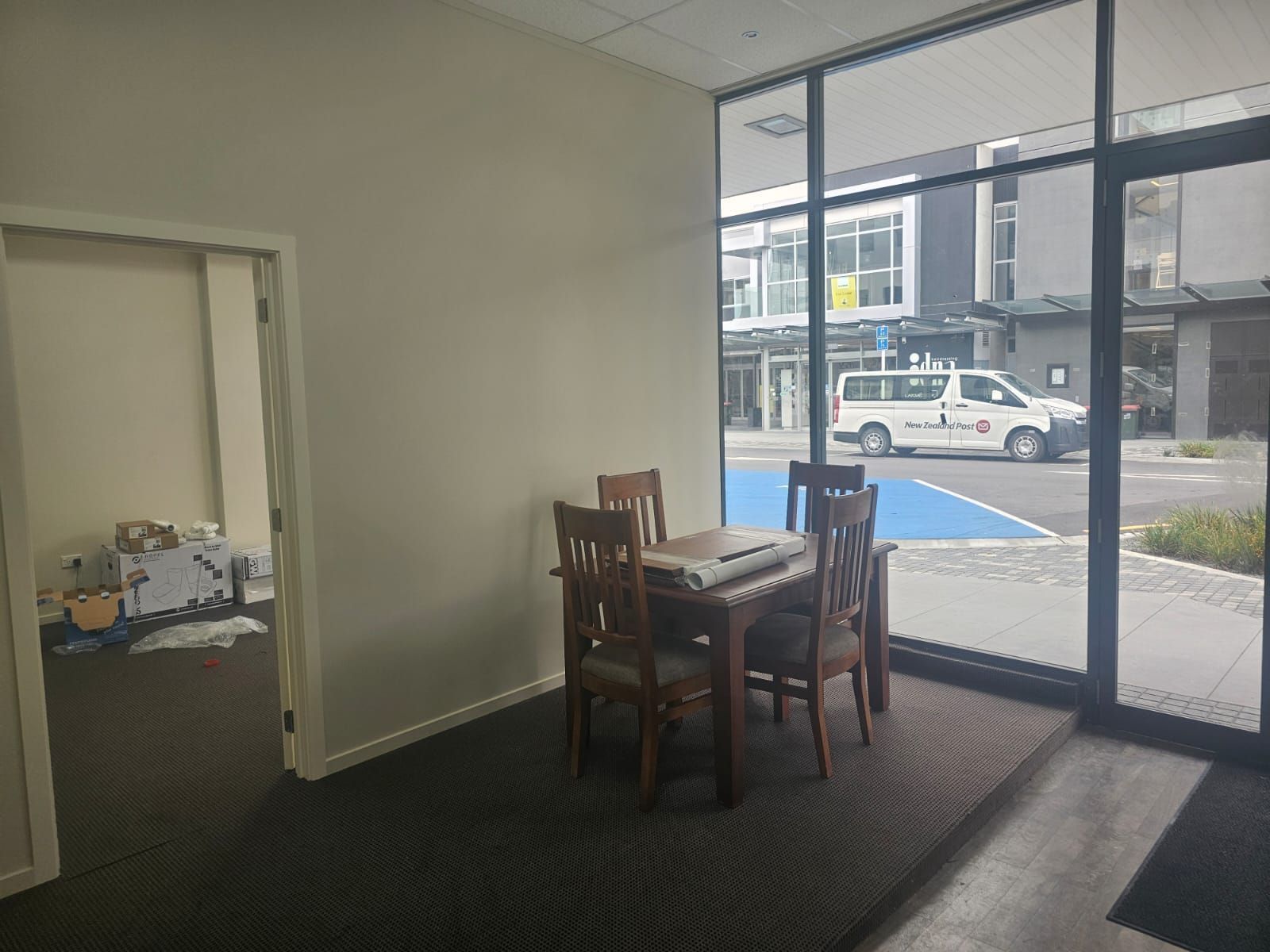 An empty room with a dining table and chairs near a large window overlooking a street with buildings and a van.