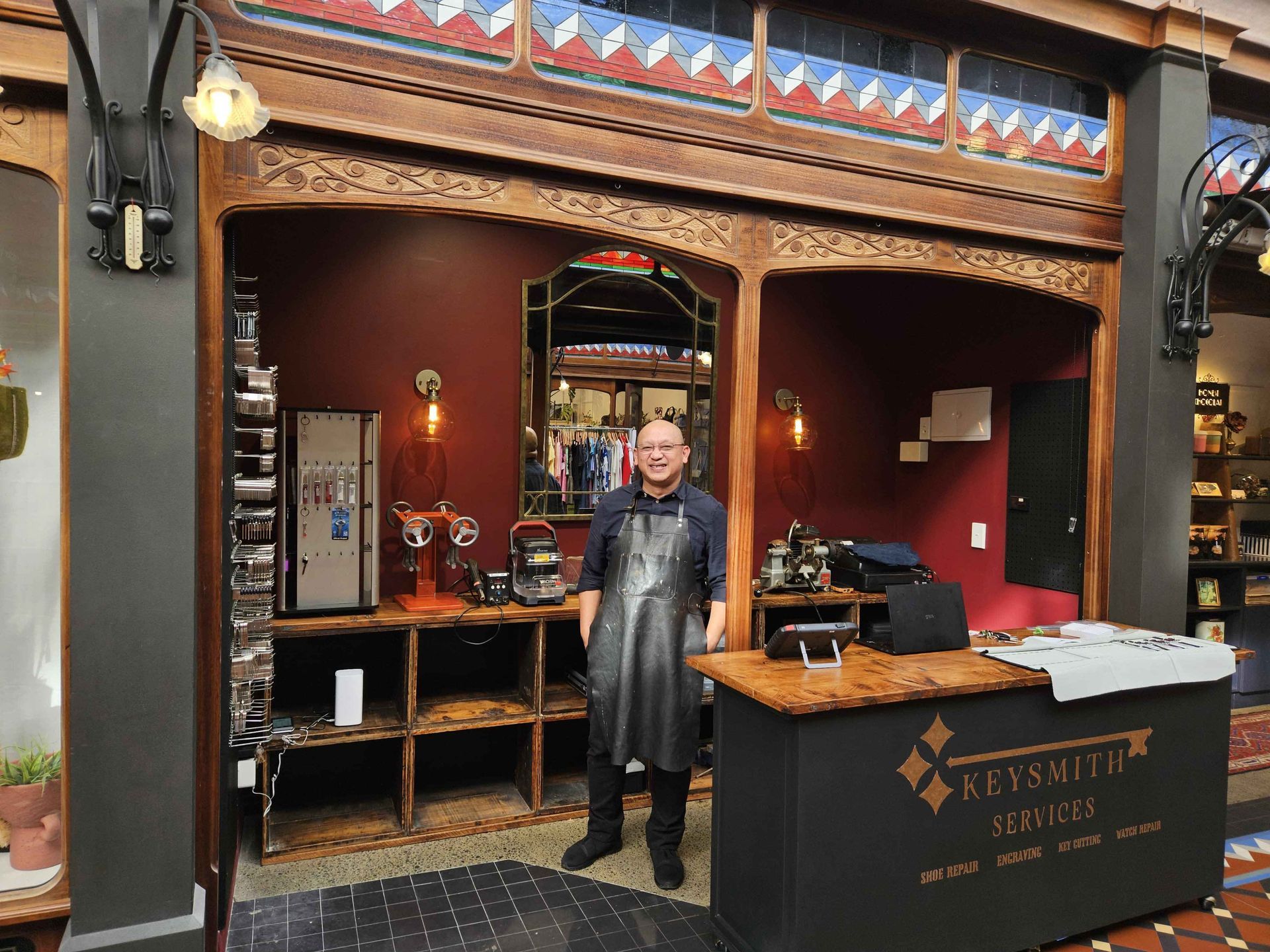 A man in a leather apron stands at the counter of a keysmith shop. Dark red walls, ornate wood trim.