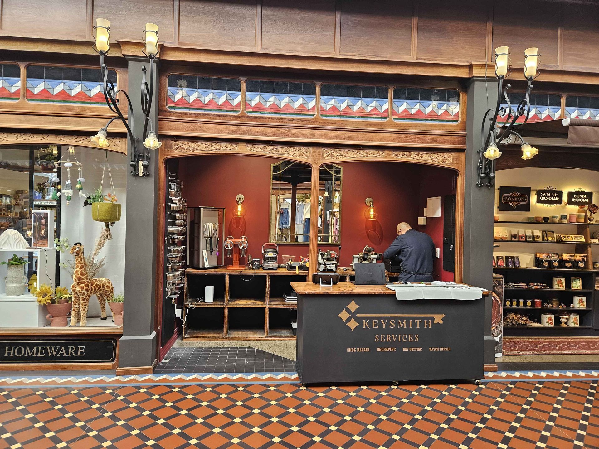 Shopfront with ornate wooden trim and shelves. A person stands behind a counter. Red and brown tones.
