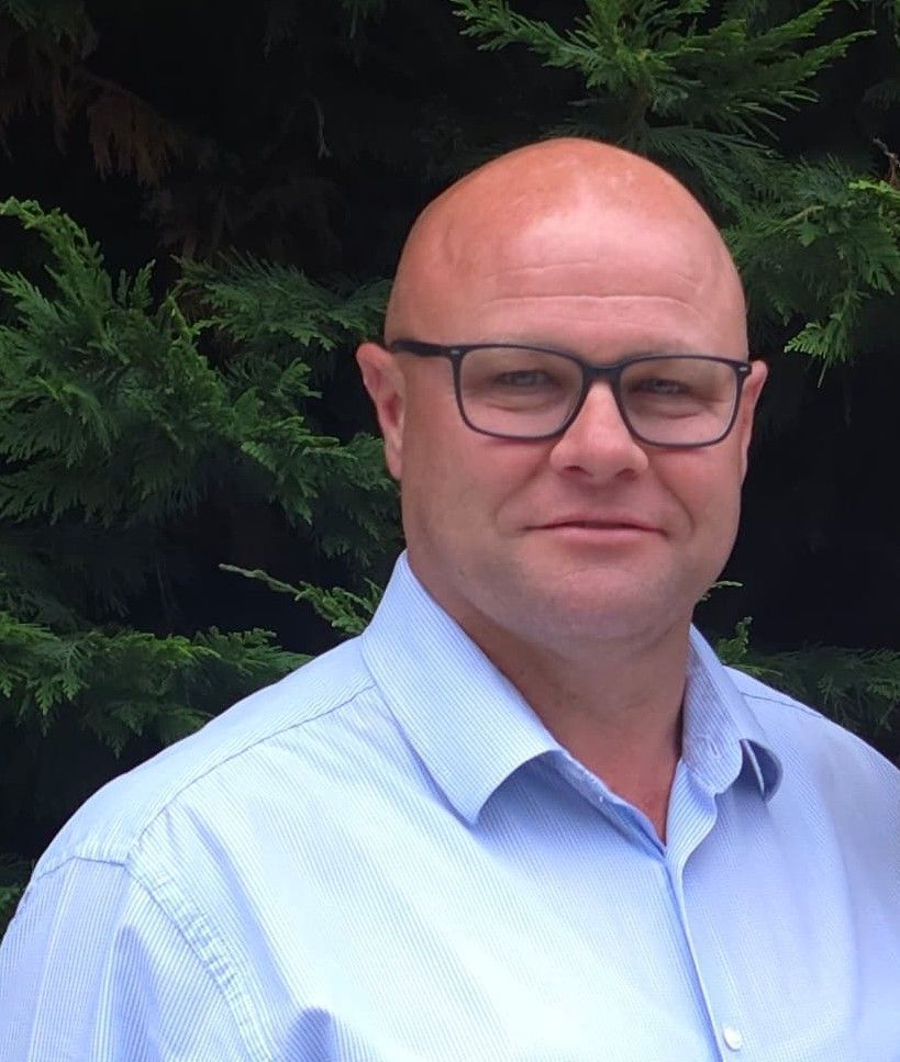 Bald man with glasses, light blue shirt, smiling against a background of green foliage.
