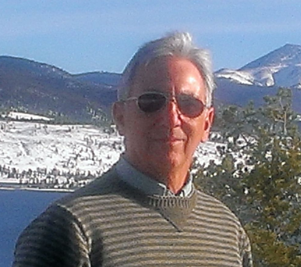 Man in sunglasses and sweater smiles, snowy mountains and lake in the background.
