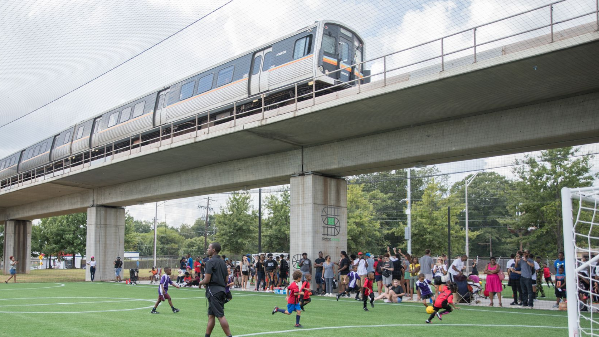 A train crosses an elevated track above a public sports field where people are gathered and children are playing soccer.