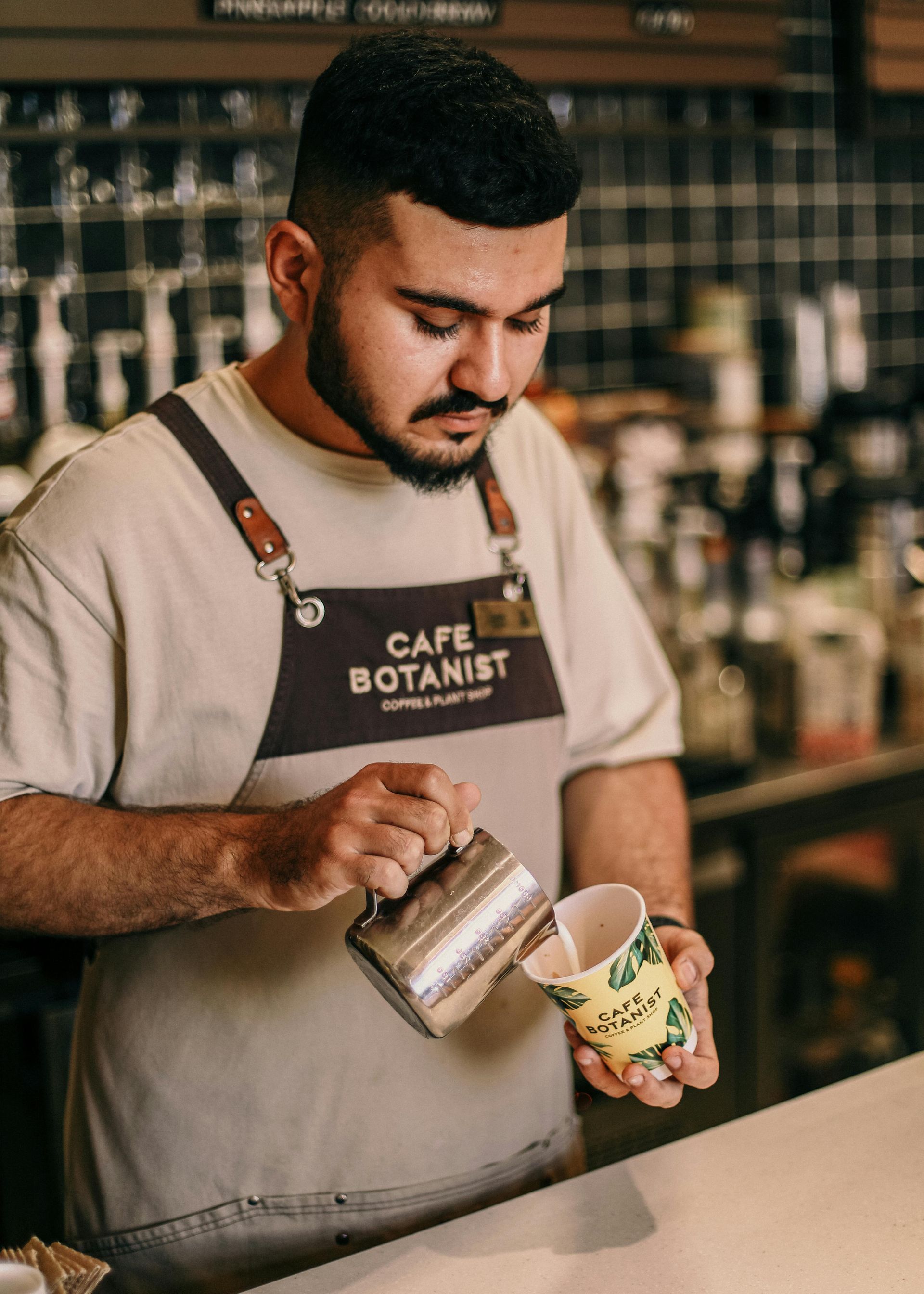 A barista in a Cafe Botanist apron pours steamed milk from a metal pitcher into a decorated paper cup.