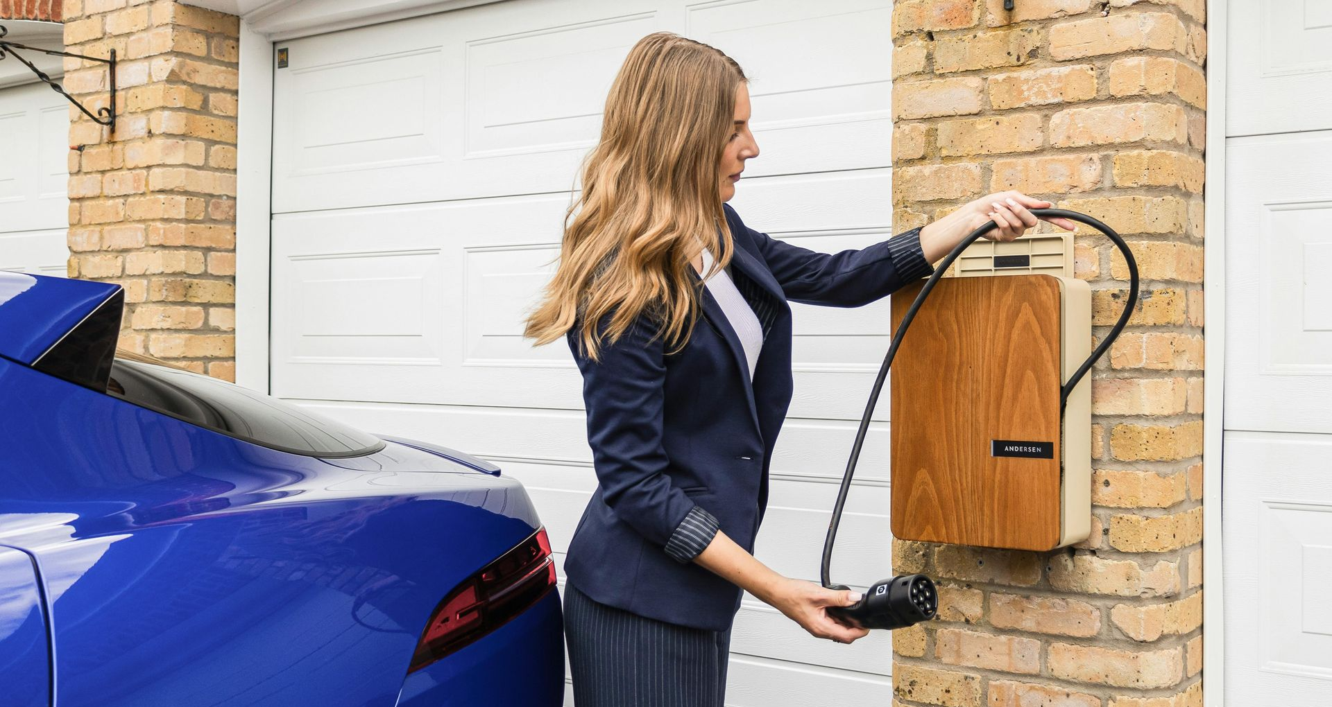 A person in business attire retrieves a charging cable from a wood-paneled wall unit to charge a blue electric car.