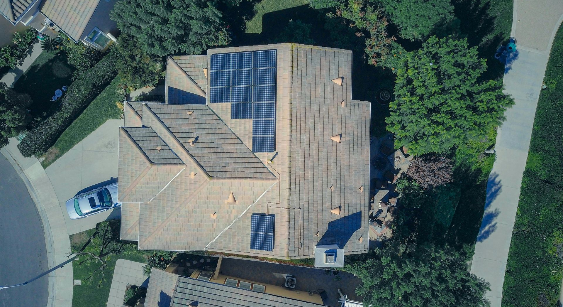Aerial view of a residential roof featuring multiple solar panel installations and a chimney, surrounded by landscaping.