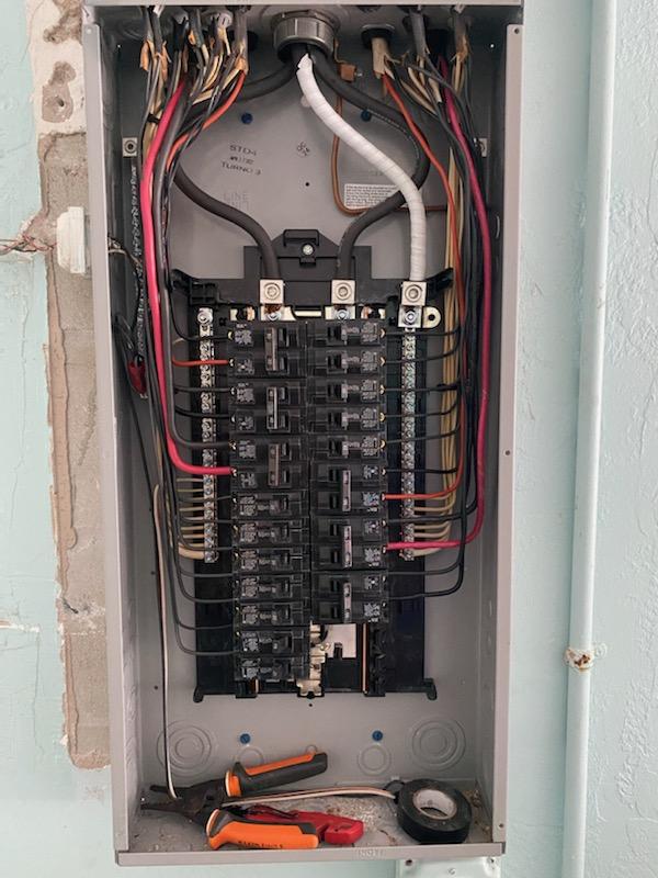 Open electrical panel with numerous black circuit breakers, wires, and tools.