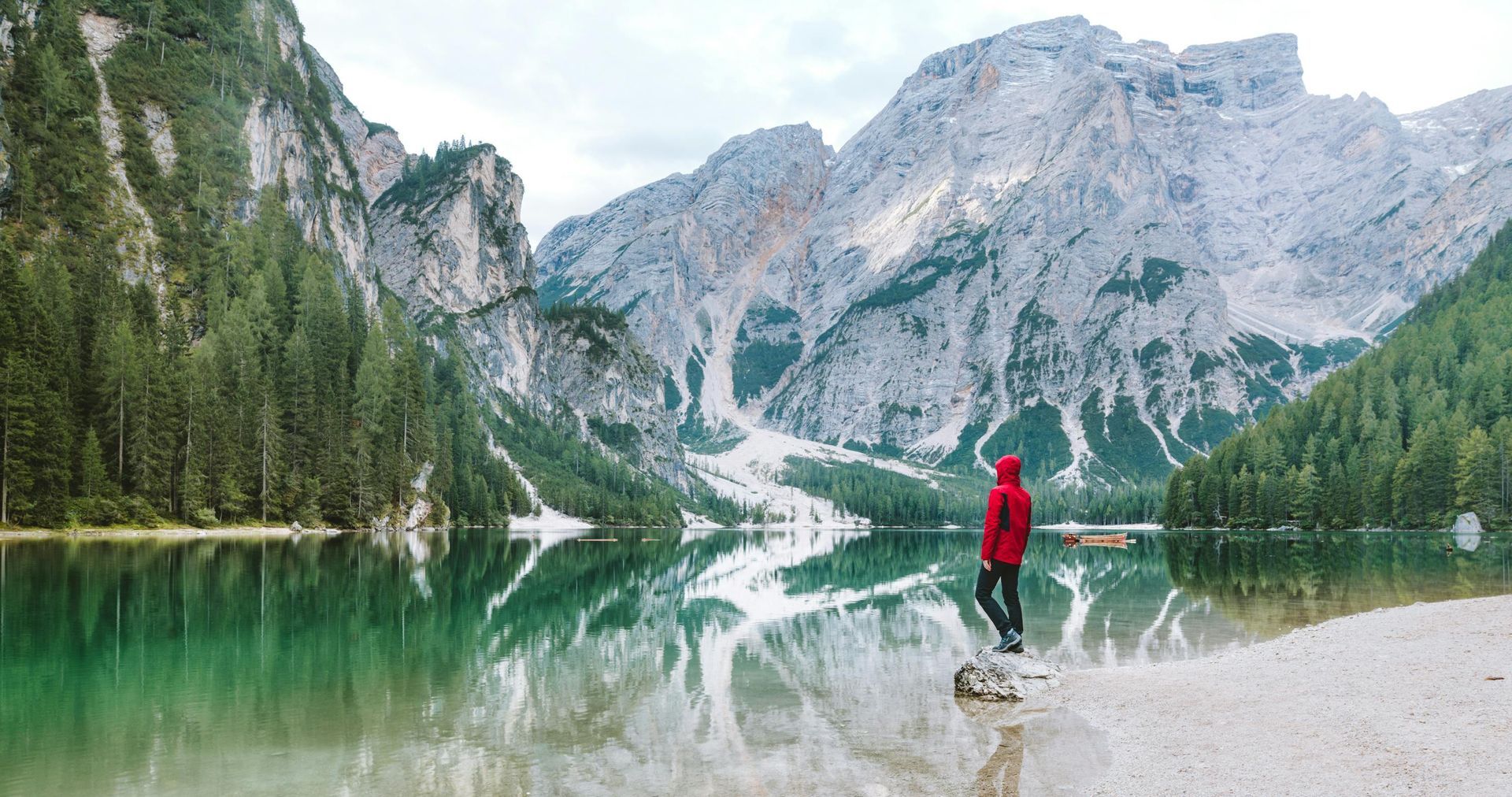 A person in a red jacket is standing on the shore of a lake in the mountains.