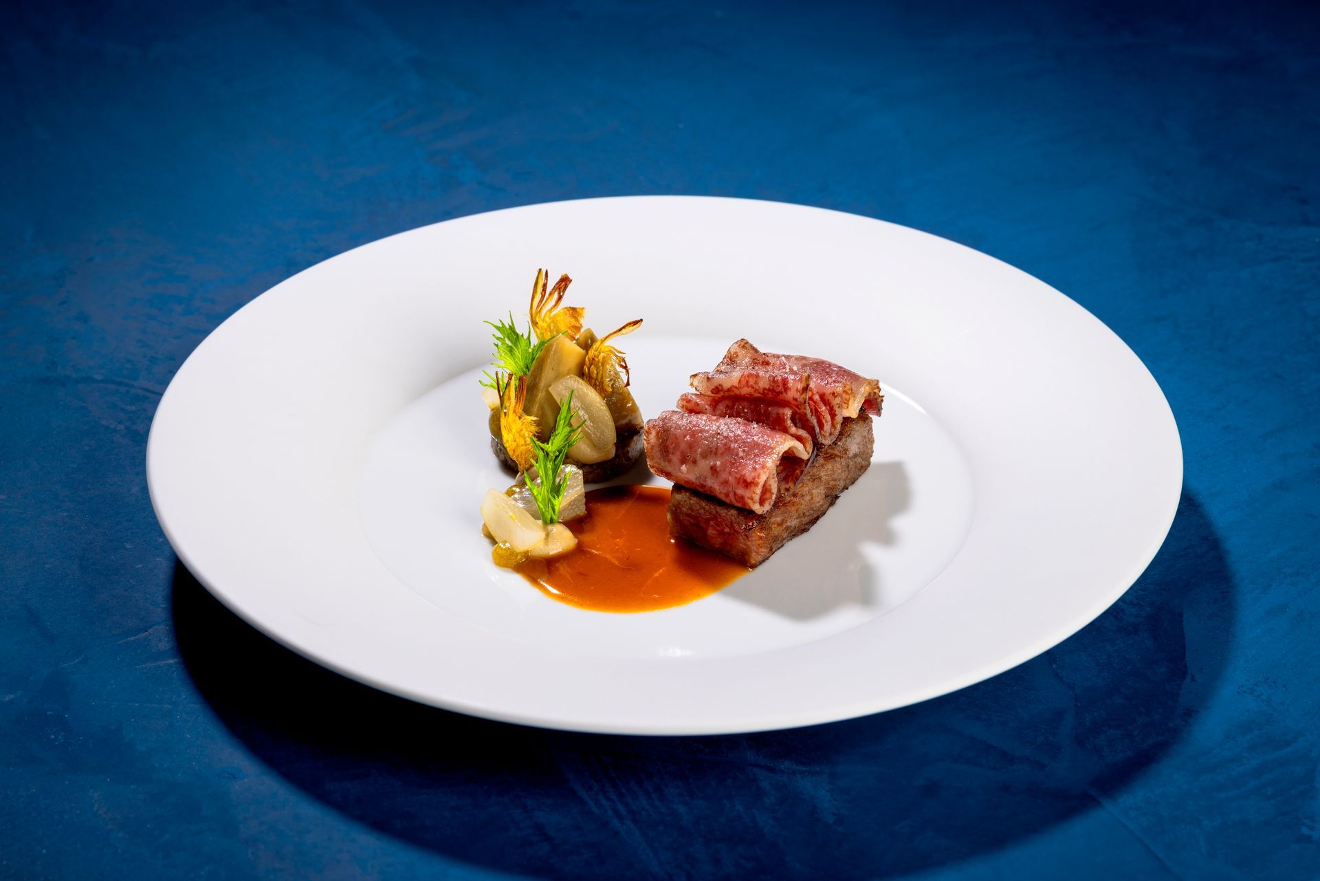 Gourmet dish on a white plate: seared meat on a dark base, sauce, and vegetable garnish on a blue background.