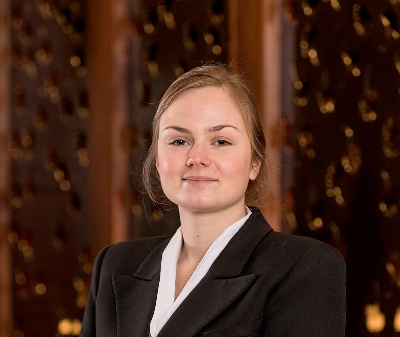 A woman in a black suit and white collar is standing in front of a wooden wall.