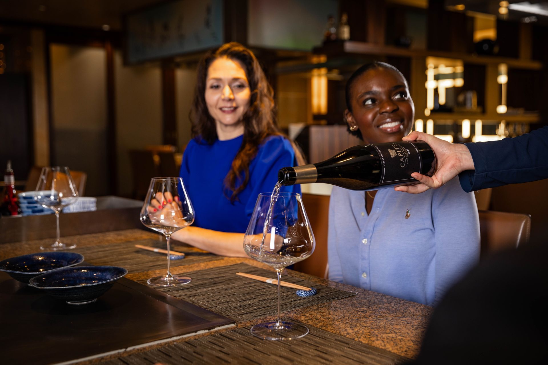 A woman is sitting at a table while a man pours wine into a glass.