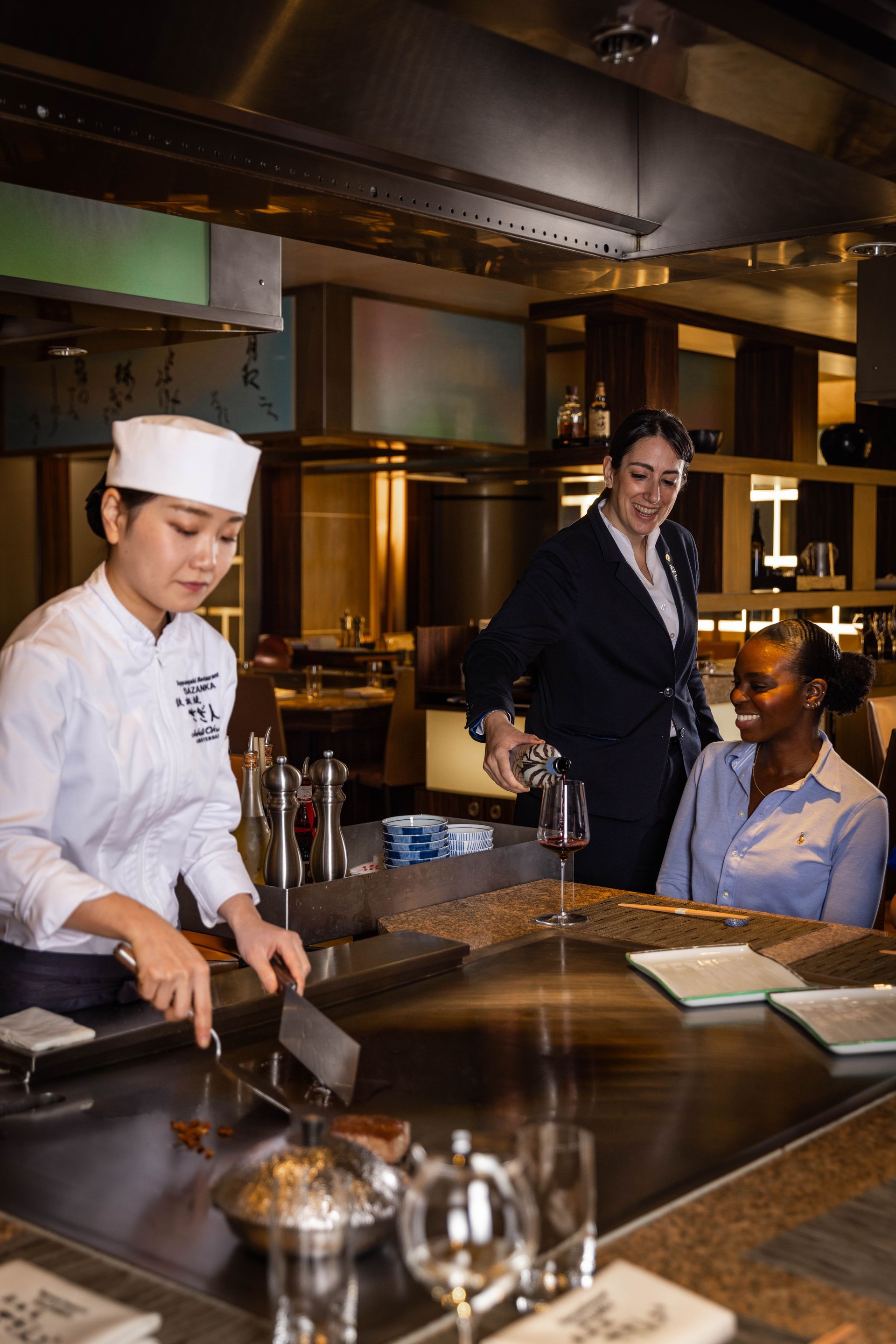 A woman is pouring wine into a glass while a chef prepares food in a kitchen.