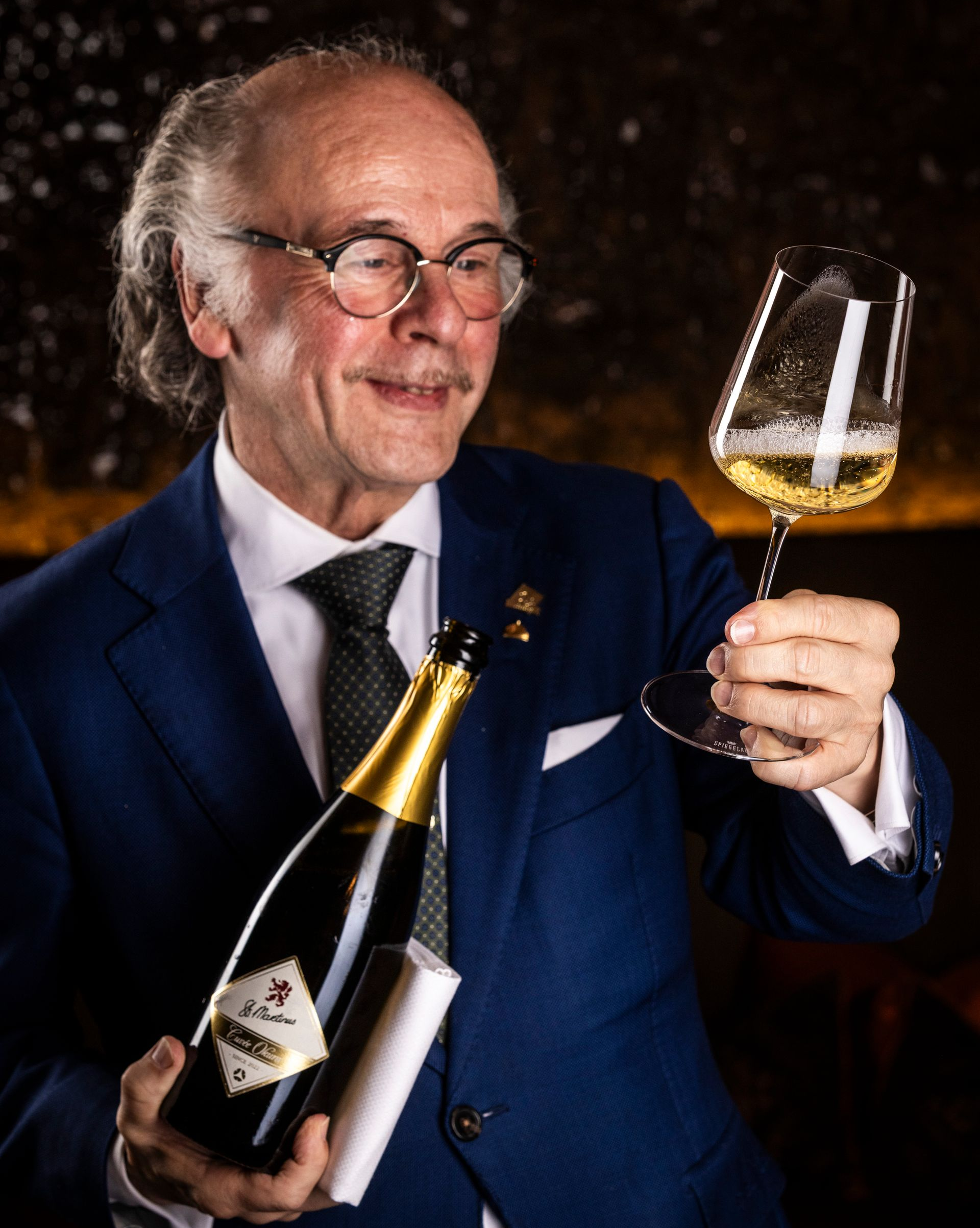Man in pak met een fles champagne en een glas, die de wijn inspecteert.