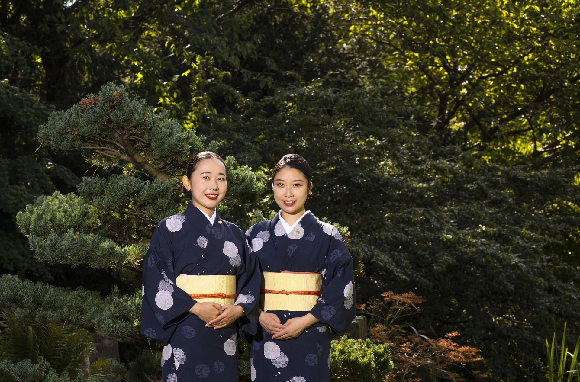 Two women in kimonos are standing next to each other in a forest.