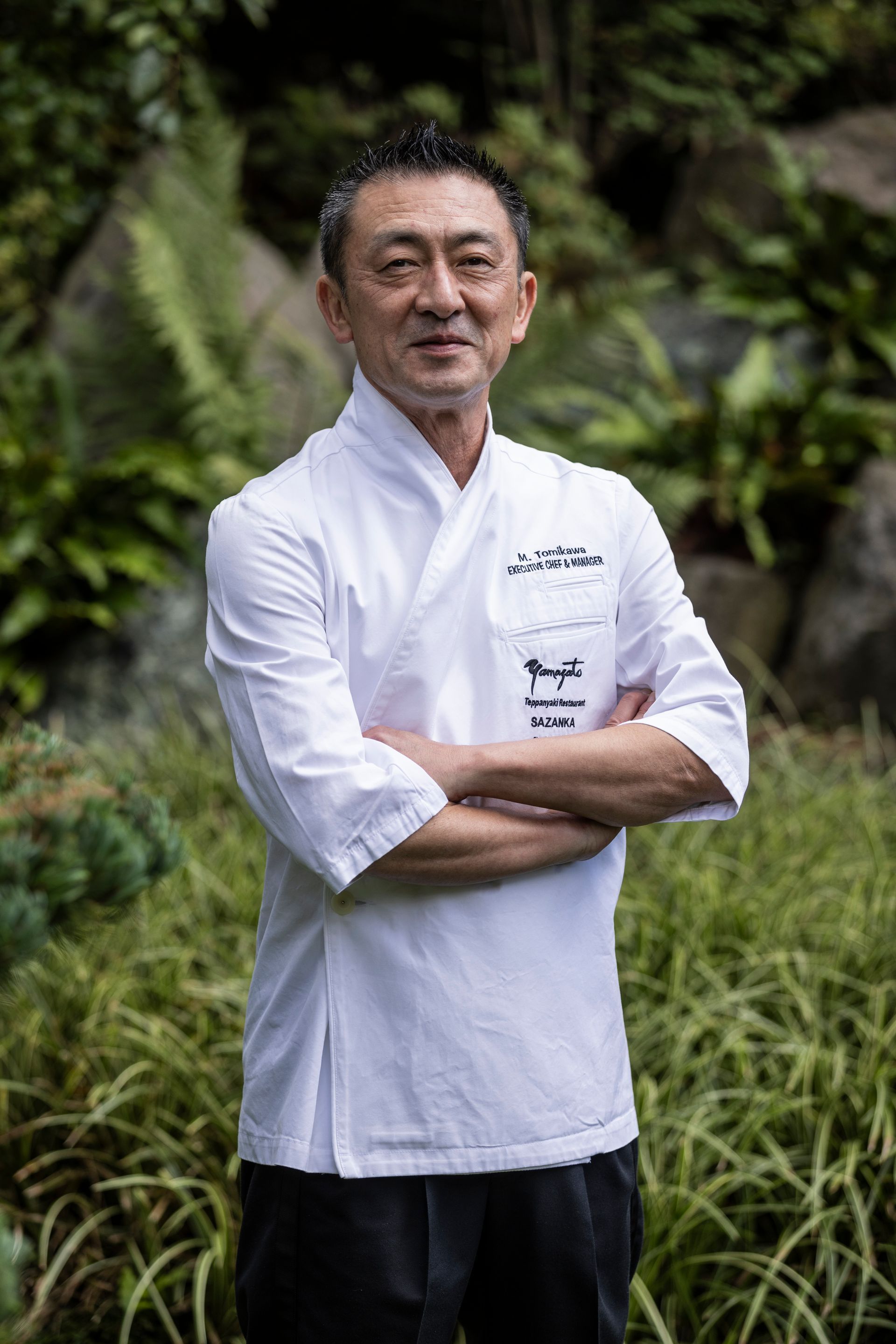A man in a white chef 's jacket is standing in a garden with his arms crossed.