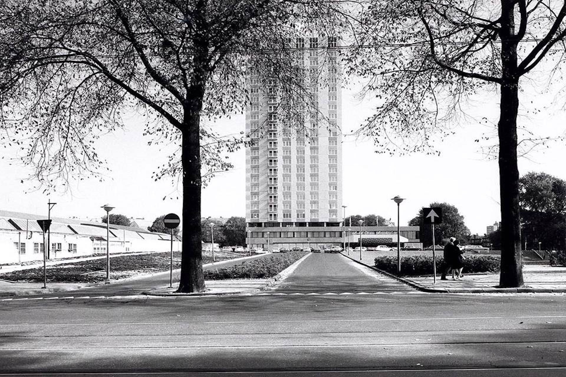 A black and white photo of a street with trees and a tall building in the background