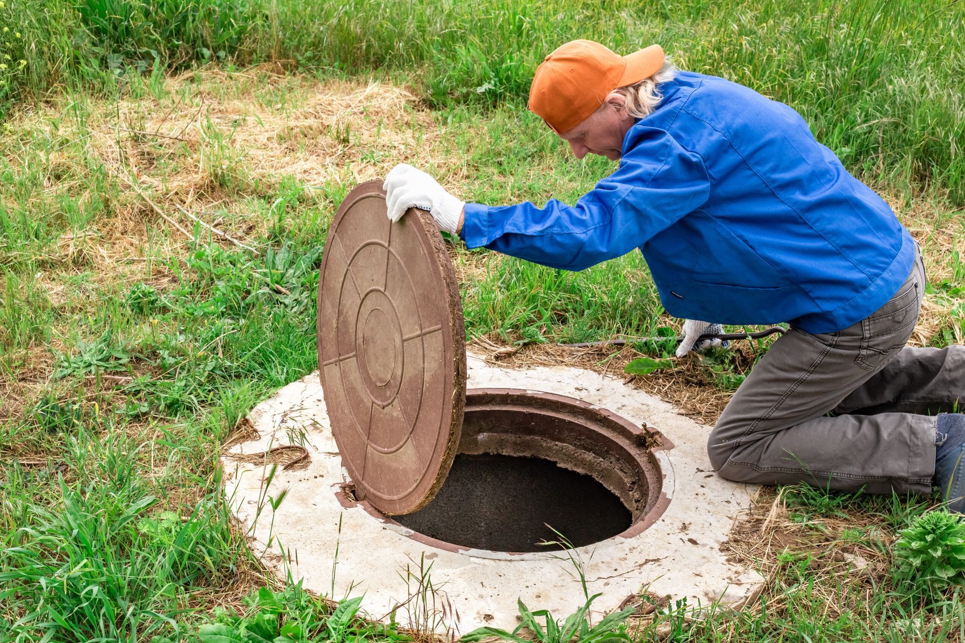 Person in blue jacket lifting a manhole cover beside a grassy ground opening