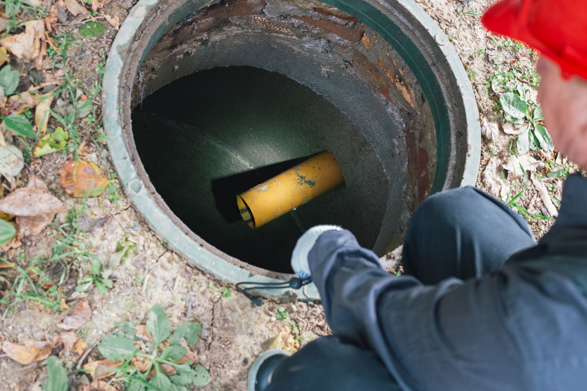 Person peering into an open manhole with a yellow flashlight inside, outdoors among leaves.