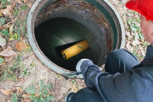 Person looking into an open manhole with a yellow pipe inside, outdoors on grass and leaves