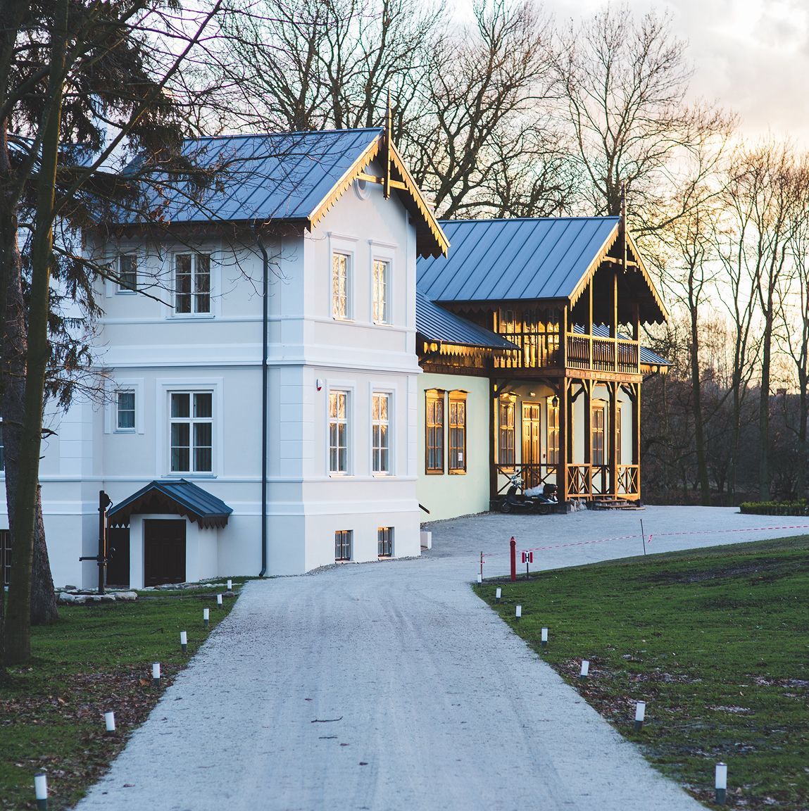 White house with blue roofs, long driveway, surrounded by trees and grass, dusk lighting.