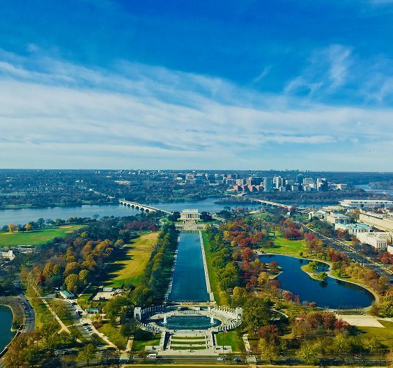 Aerial view of Washington, D.C., with reflecting pool, monuments, autumn trees, and blue sky.