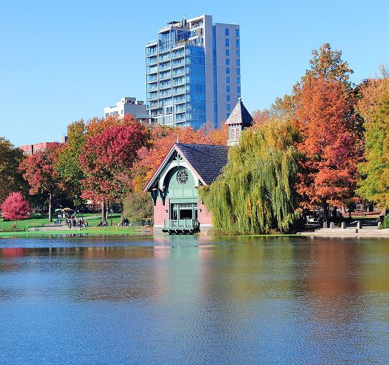 Boathouse on a lake surrounded by fall foliage, with a modern building in the background.