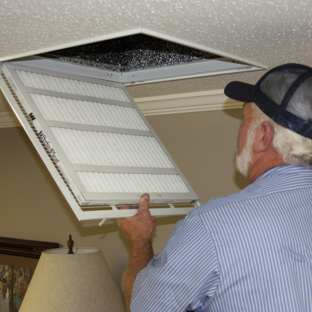 Person replacing air filter in ceiling vent. Filter is white with gray frame. Man wears blue shirt and a baseball cap.