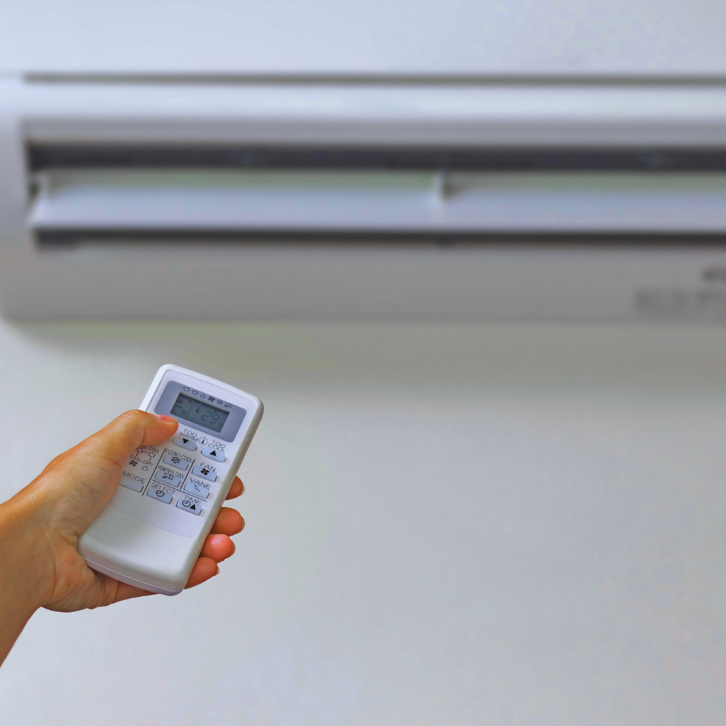 Hand holding a remote control pointing towards a white air conditioning unit on a white wall.