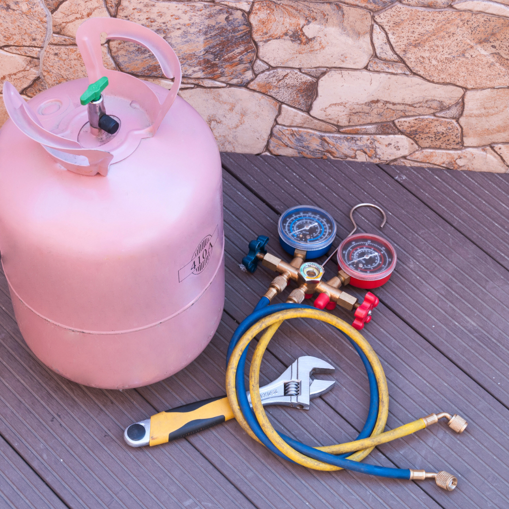 Pink refrigerant tank with gauges, hoses, wrench, and utility knife on a wooden surface.