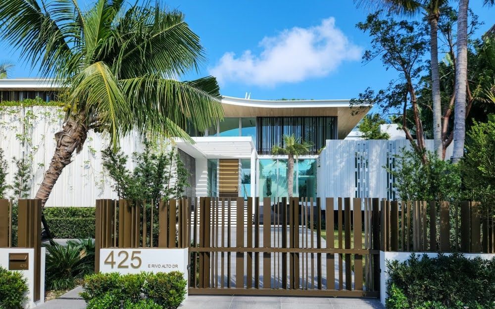 A luxurious Miami home with reflective window tint, framed by palm trees and a gated yard.