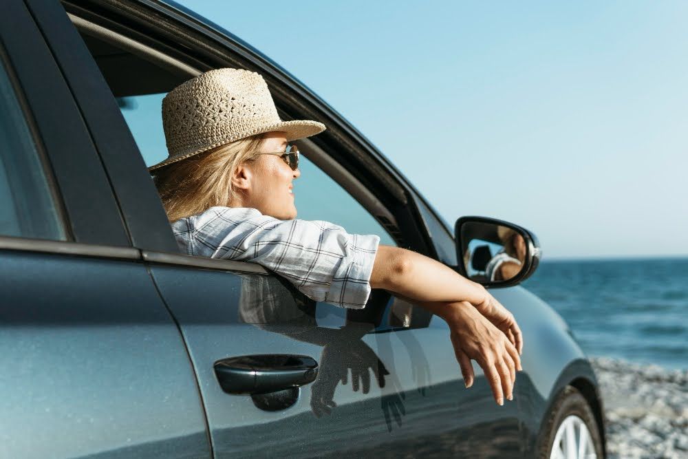A woman looking out of a car window with the sea in the background on a sunny day.