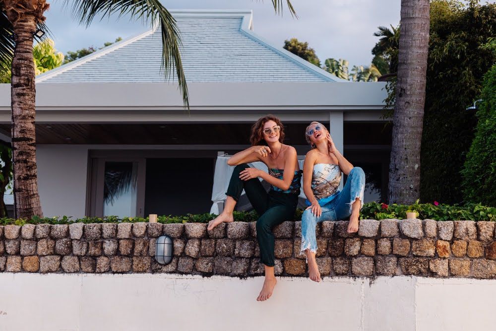 Two women sitting on a rock fence outside a house with tinted windows.