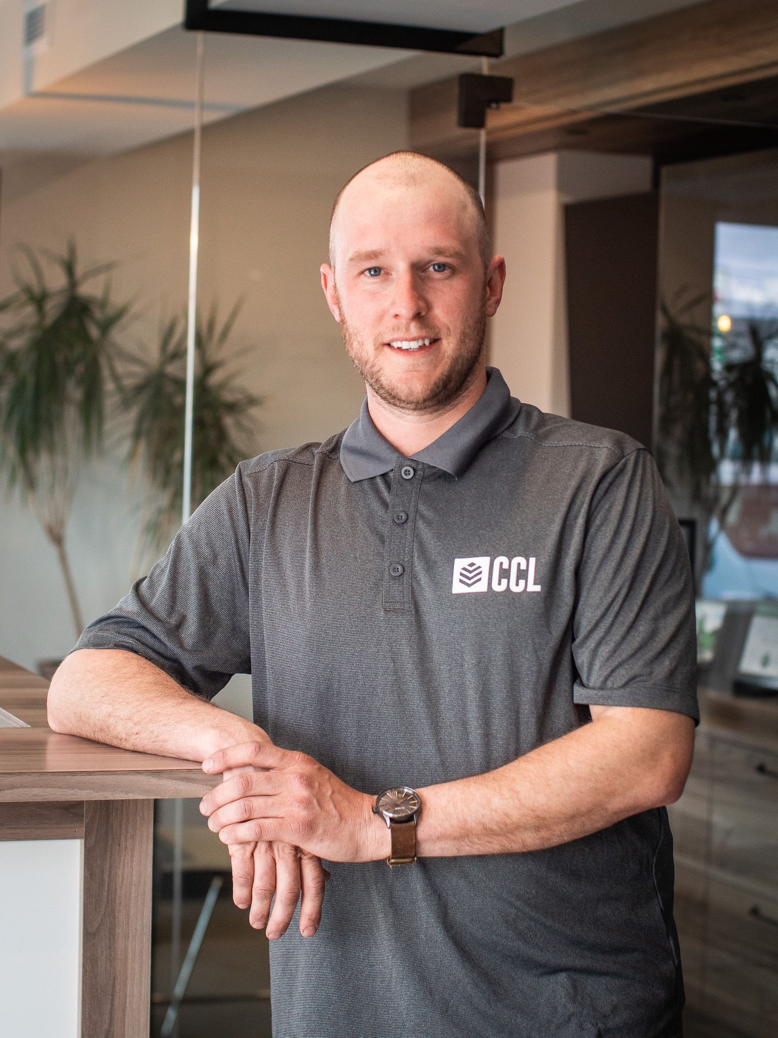 Un homme en polo gris s'appuie sur une table en bois.