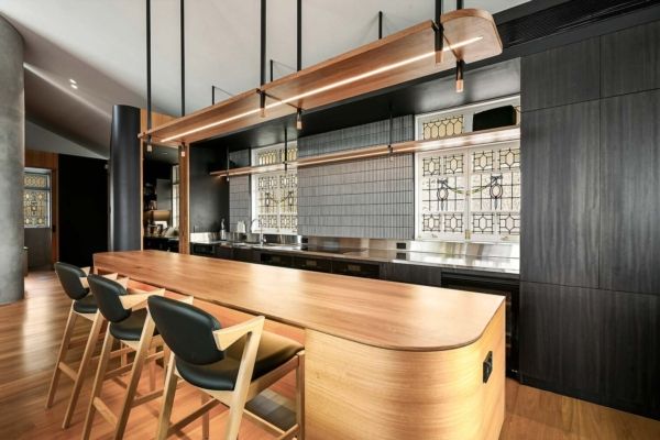 A modern bar with light wood counter and black stools, set against dark wood paneling and a patterned tile backsplash — PK Kitchen Design in West Ipswich, QLD